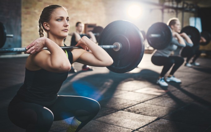 Fit young woman lifting barbells looking focused, working out in a gym with other people